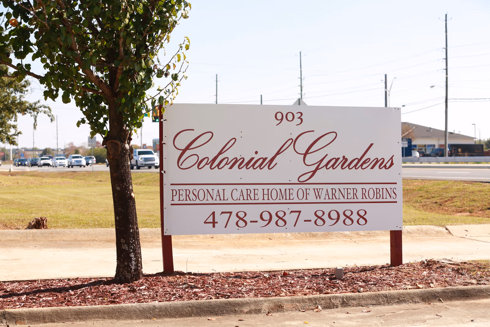 Outdoor view of a white sign with maroon text that reads '903 Colonial Gardens Personal Care Home of Warner Robins 478-987-8988' next to a tree and a road with cars in the background.