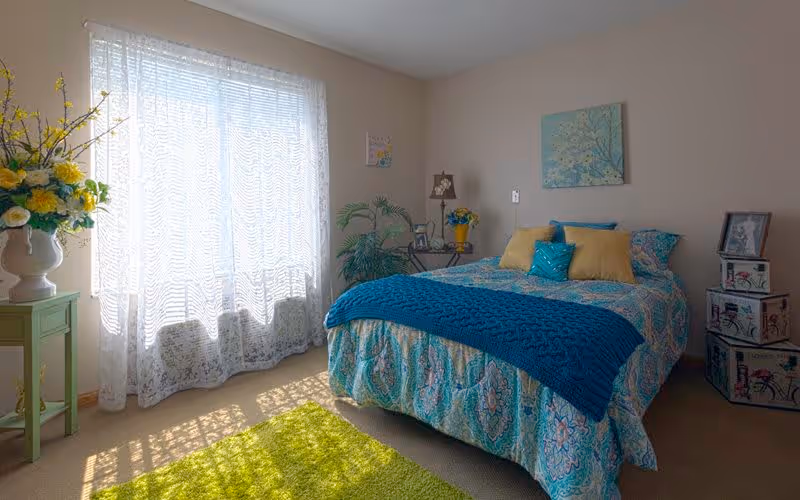 A bright bedroom with a large window covered by sheer white curtains allowing sunlight to fill the room. The bed is made with a colorful patterned bedspread in shades of blue and teal, accented with yellow and teal pillows and a blue knitted throw blanket. There is a green rug on the floor, a small green side table with a vase of yellow and white flowers, a potted plant in the corner, and decorative boxes stacked near the bed. The walls are light-colored with framed artwork and a lamp on a small table beside the bed.
