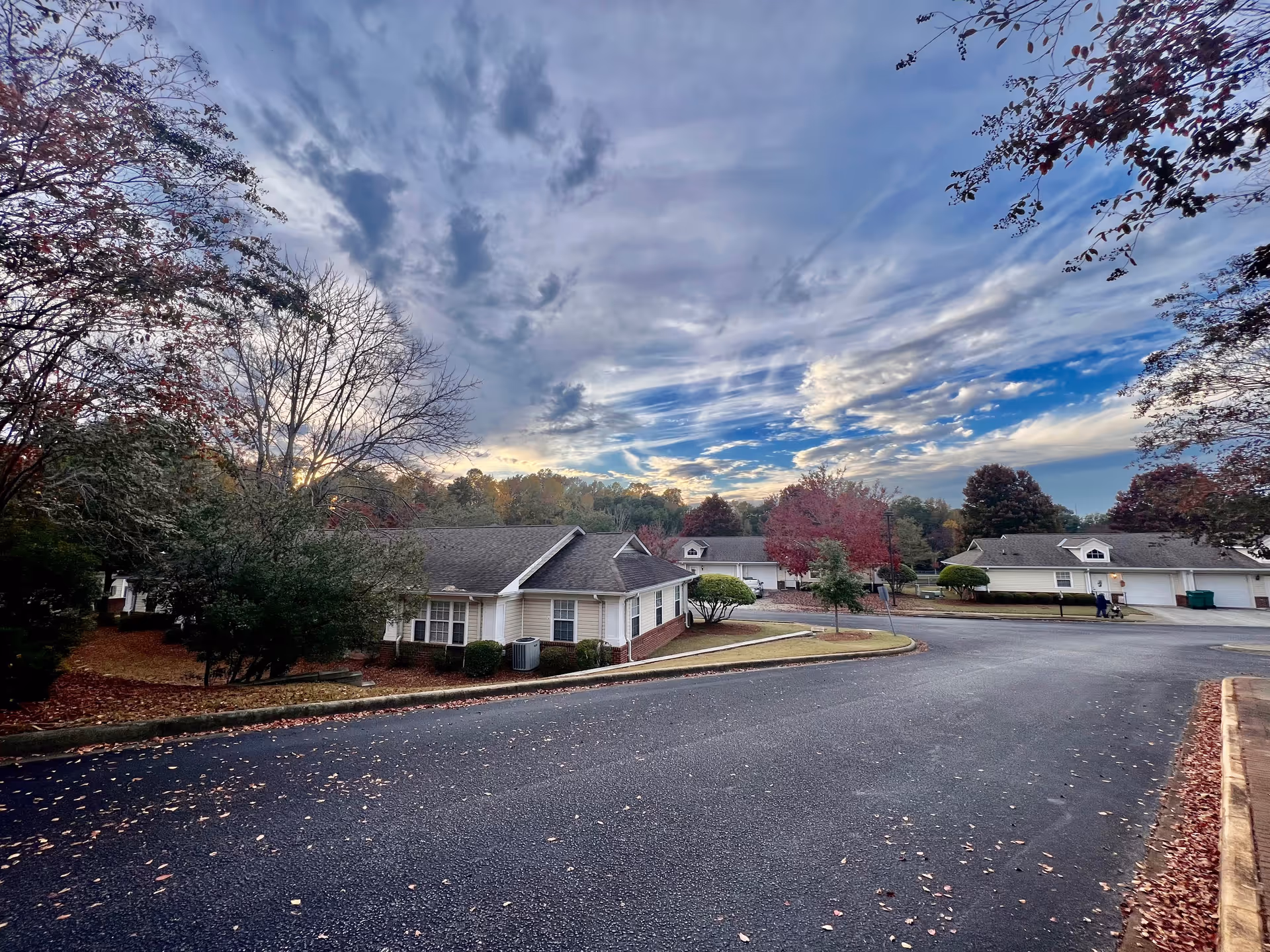 A quiet residential street in a senior living community with single-story houses surrounded by trees with autumn foliage under a partly cloudy sky during sunset.
