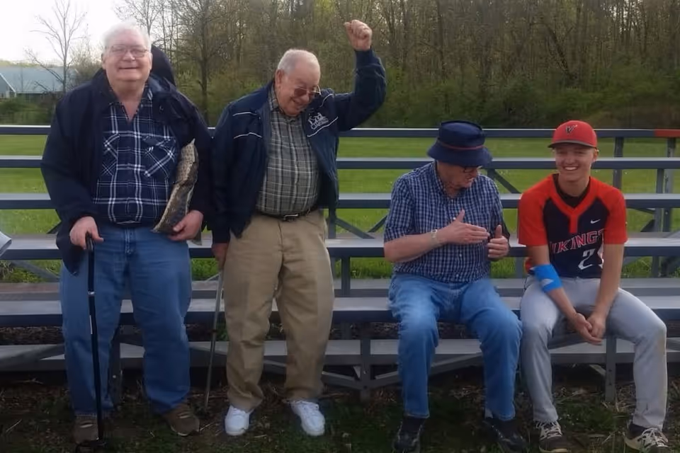 Four people sitting and standing on metal bleachers outdoors. Three elderly men, two standing and one sitting, are dressed casually with jackets and hats. A younger person in a red and black baseball uniform is sitting on the bleachers smiling. Trees and grass are visible in the background.