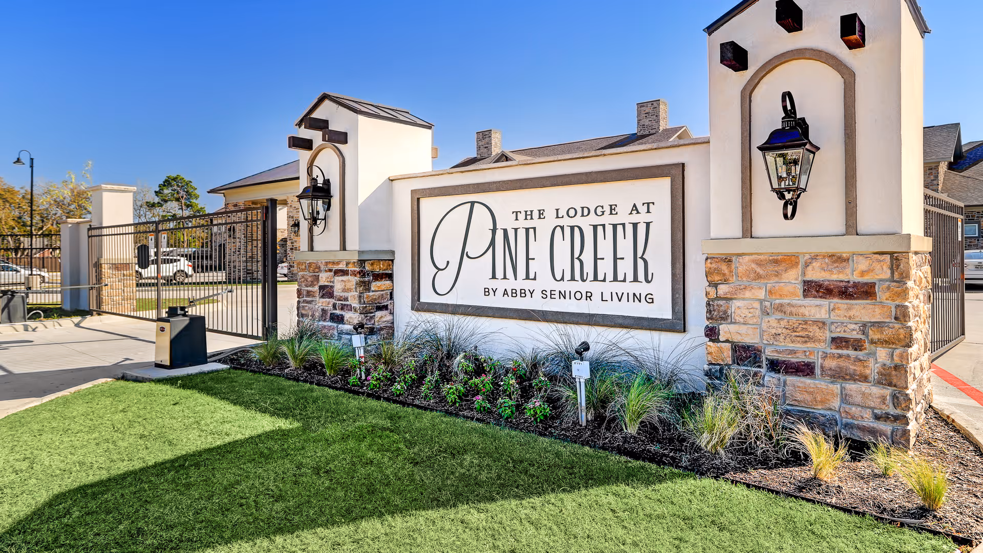 Entrance sign for The Lodge at Pine Creek by Abby Senior Living, featuring a stone and stucco structure with decorative lanterns on either side, a gated driveway, and landscaped greenery in front under a clear blue sky.