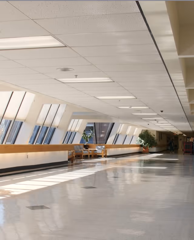A long, bright hallway with large slanted windows on one side letting in natural light. There are wooden benches with cushions and potted plants along the wall. The ceiling has recessed lighting panels, and the floor is shiny and reflective.