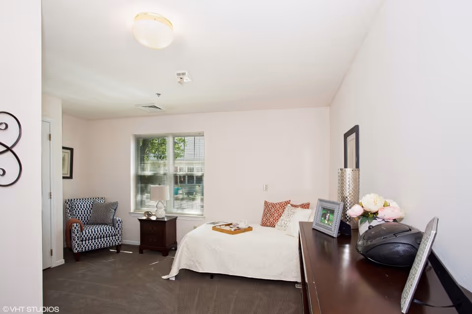 A bright bedroom with a single bed covered in a white quilt and decorative pillows. Next to the bed is a wooden nightstand with a lamp and a small decorative item. A patterned armchair with a throw blanket and pillow is placed near a window with blinds. A dark wooden dresser holds framed photos, a vase with flowers, and a radio. The walls are white, and the carpet is gray.