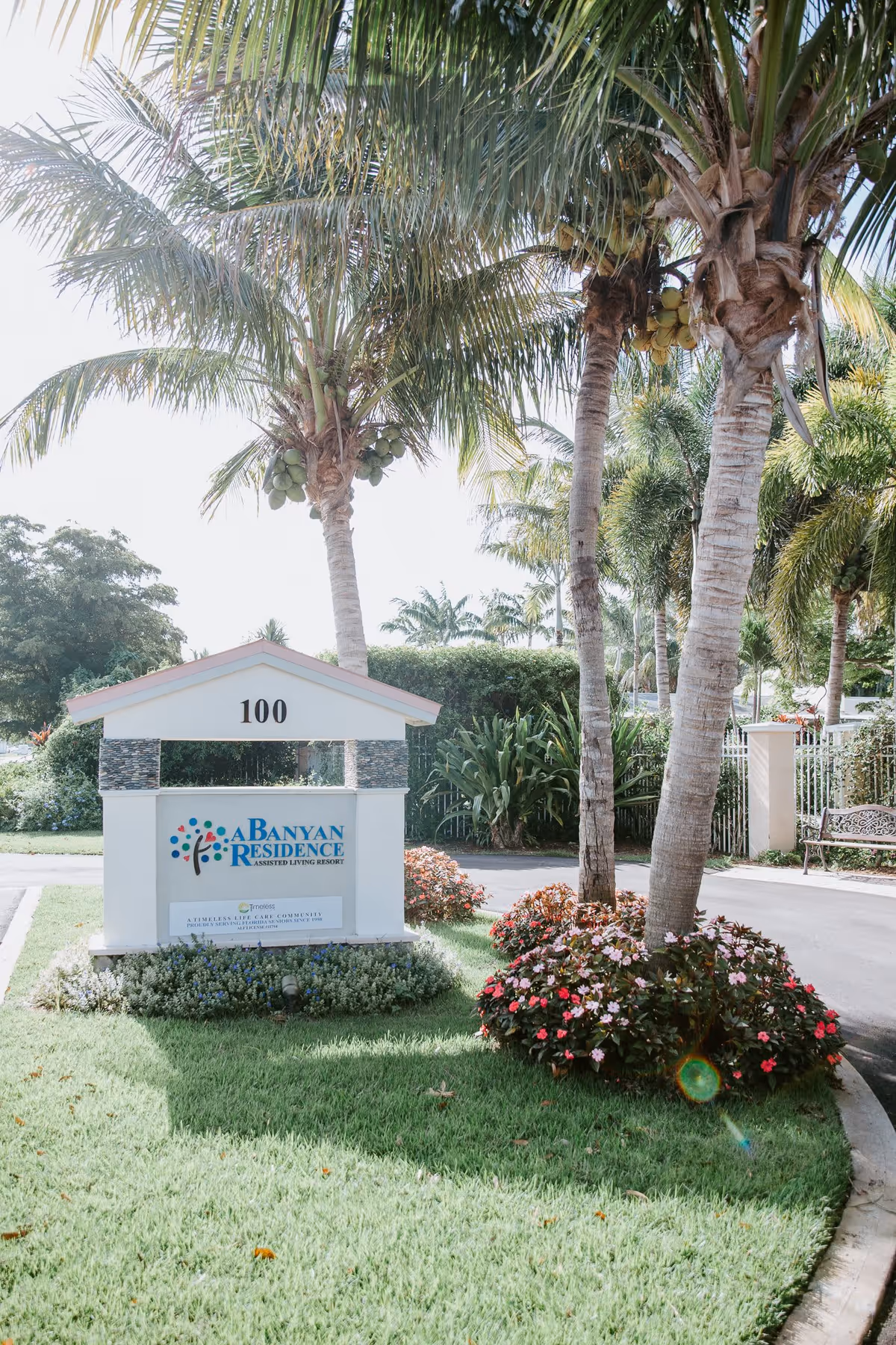 Outdoor view of the entrance sign for A Banyan Residence assisted living resort, surrounded by palm trees, green grass, and colorful flowering plants.