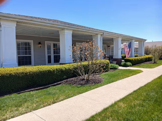 Exterior view of a single-story building with white columns and a covered porch. There are trimmed bushes and a small tree in front, a concrete walkway, and an American flag displayed near the entrance under a clear blue sky.