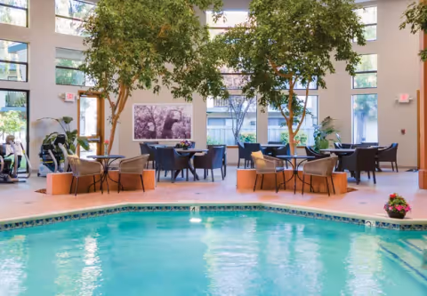 Indoor pool with turquoise water in a communal lounge area featuring tables, chairs, potted trees, and large windows.