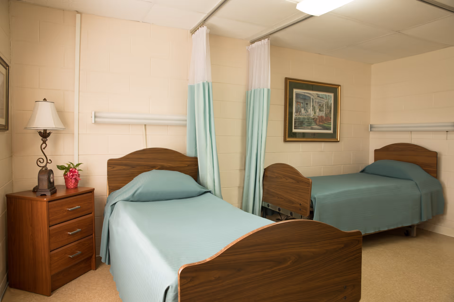 A room with two single beds separated by a curtain. Each bed has a wooden headboard and footboard, and is covered with a light blue bedspread. There is a wooden nightstand with three drawers between the beds, topped with a lamp and a small potted plant. A framed picture hangs on the beige brick wall above the bed on the right.