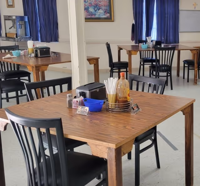A dining area with wooden tables and black chairs. Each table has condiments, napkin holders, and utensils. Blue curtains cover the windows, and a framed painting hangs on the wall.