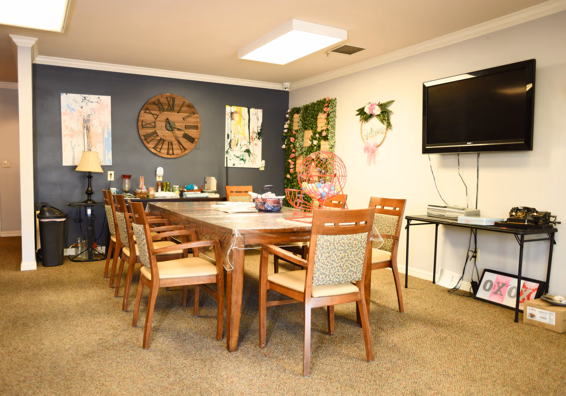 A well-lit common room with a large wooden table surrounded by eight wooden chairs with cushioned seats and backs. The room features a dark accent wall with two abstract paintings and a large wooden clock. On the opposite wall, there is a flat-screen TV mounted above a black table holding electronic devices and decorative items. The room is carpeted and decorated with a green floral wall hanging and a wreath with pink flowers.