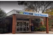 Entrance of The New Jewish Home, Sarah Neuman facility with a covered walkway supported by brick pillars, glass doors, and surrounding trees with autumn foliage.