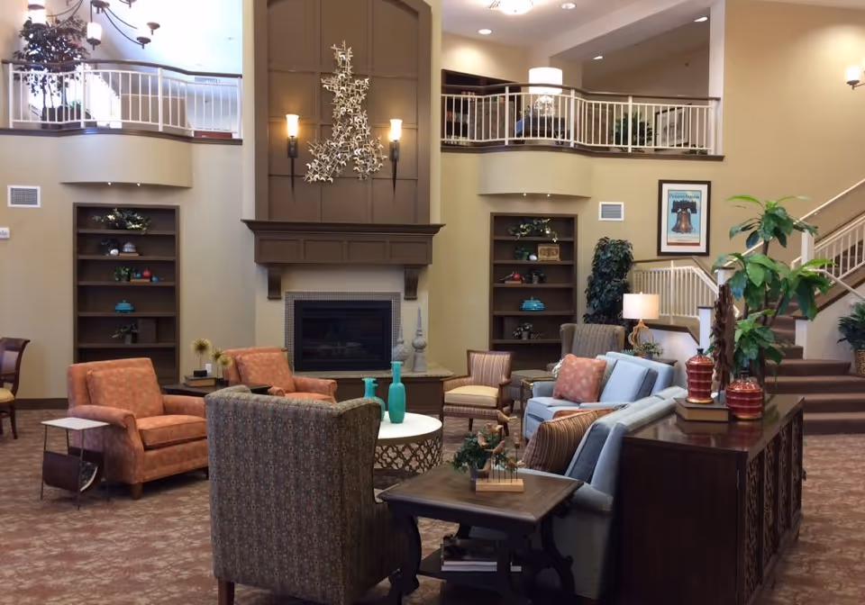 Lobby-style common room with sofas and armchairs arranged around a fireplace beneath an upstairs balcony.