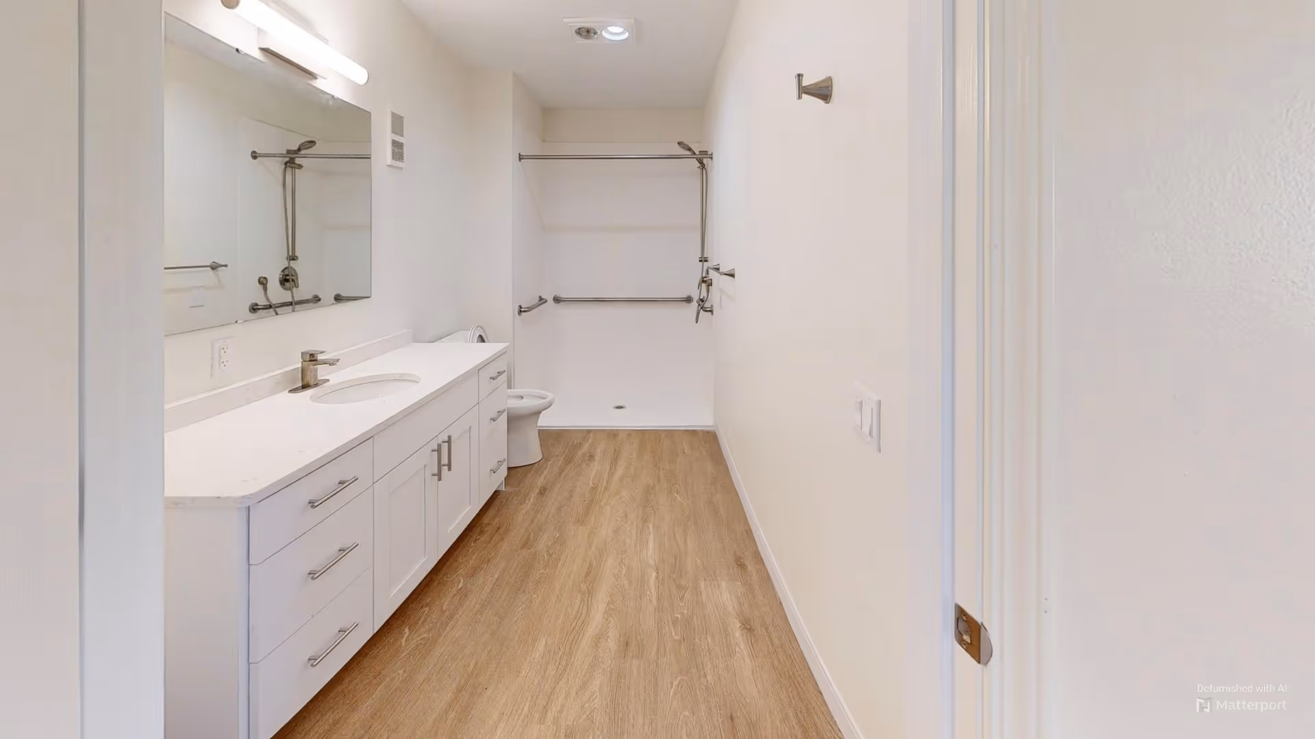A modern bathroom with a long white vanity featuring multiple drawers and a sink, a large mirror above the vanity, a toilet, and a walk-in shower area with grab bars and a handheld showerhead. The floor is covered with wood-like flooring and the walls are painted white.