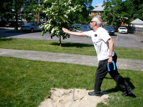 An elderly man wearing sunglasses and a white t-shirt is playing horseshoes outdoors on a grassy area near a parking lot with several cars and trees in the background.