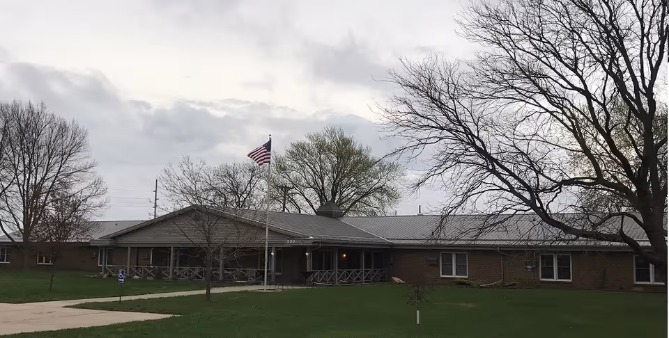 Single-story brick senior care facility with a front entrance, American flag, and leafless trees on the lawn.