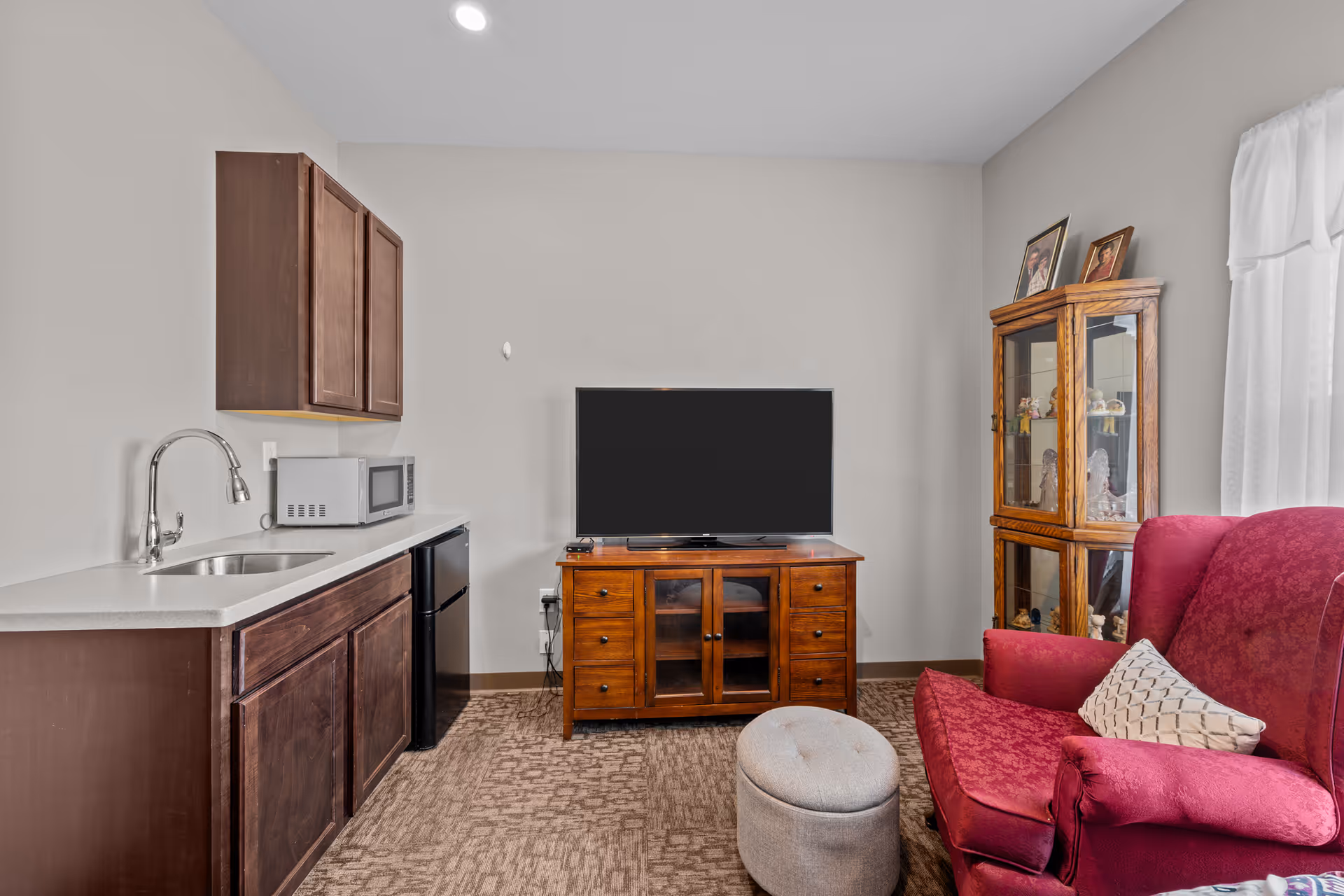 A cozy living area in a senior living facility featuring a red upholstered armchair with a patterned pillow, a small gray ottoman, a wooden TV stand with a flat-screen television, a wooden display cabinet with framed photos and figurines, and a kitchenette with a sink, microwave, mini fridge, and wooden cabinets.