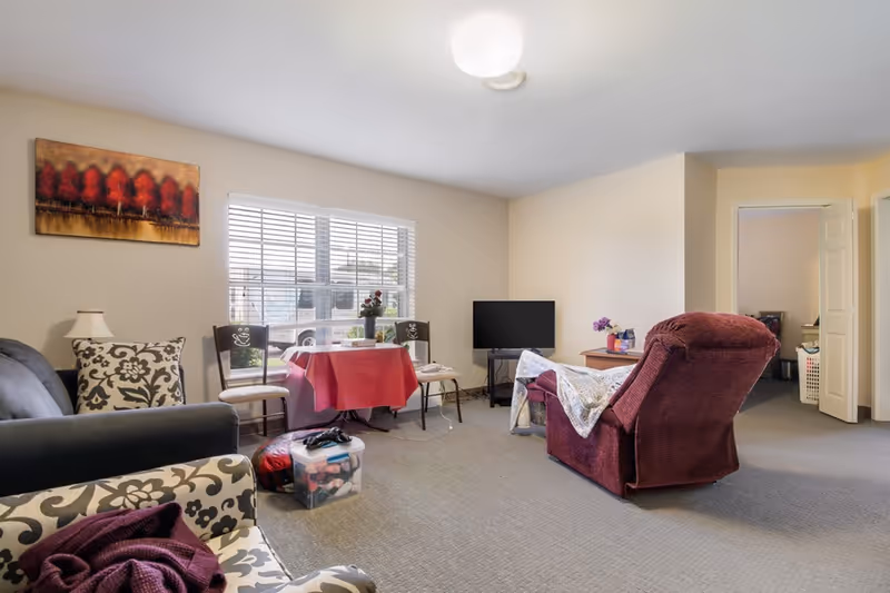 A cozy living room in a senior living facility with a large window letting in natural light. The room features a dark gray sofa with patterned pillows, a red recliner chair with a blanket draped over it, a small table covered with a red tablecloth and two chairs, a flat-screen TV on a stand, and a painting of red trees on the wall. The carpeted floor has a plastic storage container and some personal items near the sofa. An open doorway leads to another room.