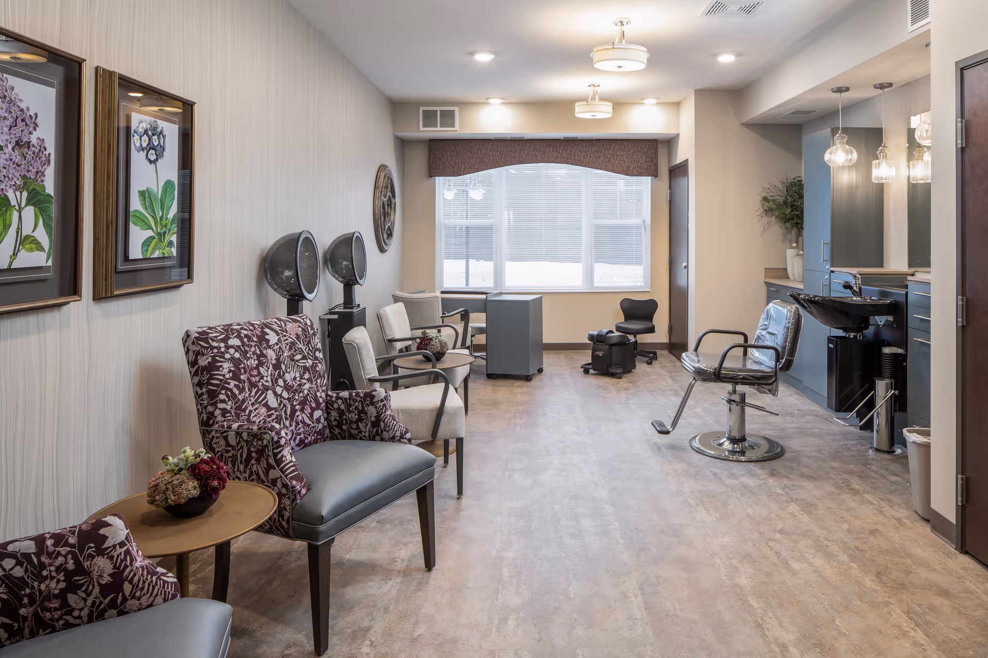 A bright and clean salon area in a senior living facility featuring floral patterned chairs, beige armchairs, hair drying stations, a styling chair, a hair washing sink, and decorative wall art. The room has large windows with blinds and soft overhead lighting.
