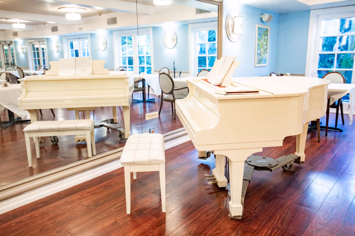 A white grand piano with a matching padded bench sits in a bright dining/activity room with tables, chairs, mirrors, and windows.