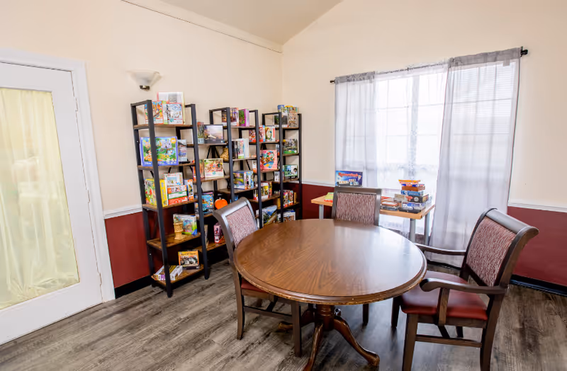 A bright room with a round wooden table surrounded by four chairs with red cushions. Against the wall, there are three black shelves filled with board games and puzzles. A small table near a window with sheer curtains also holds several board games. The room has light-colored walls with a dark red lower section and wood-look flooring.