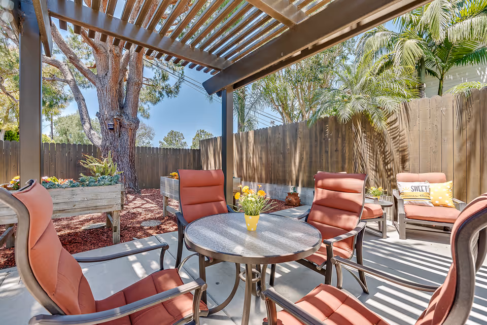 Shaded outdoor patio with a round table, cushioned chairs, potted flowers, and a pergola next to a wooden fence.