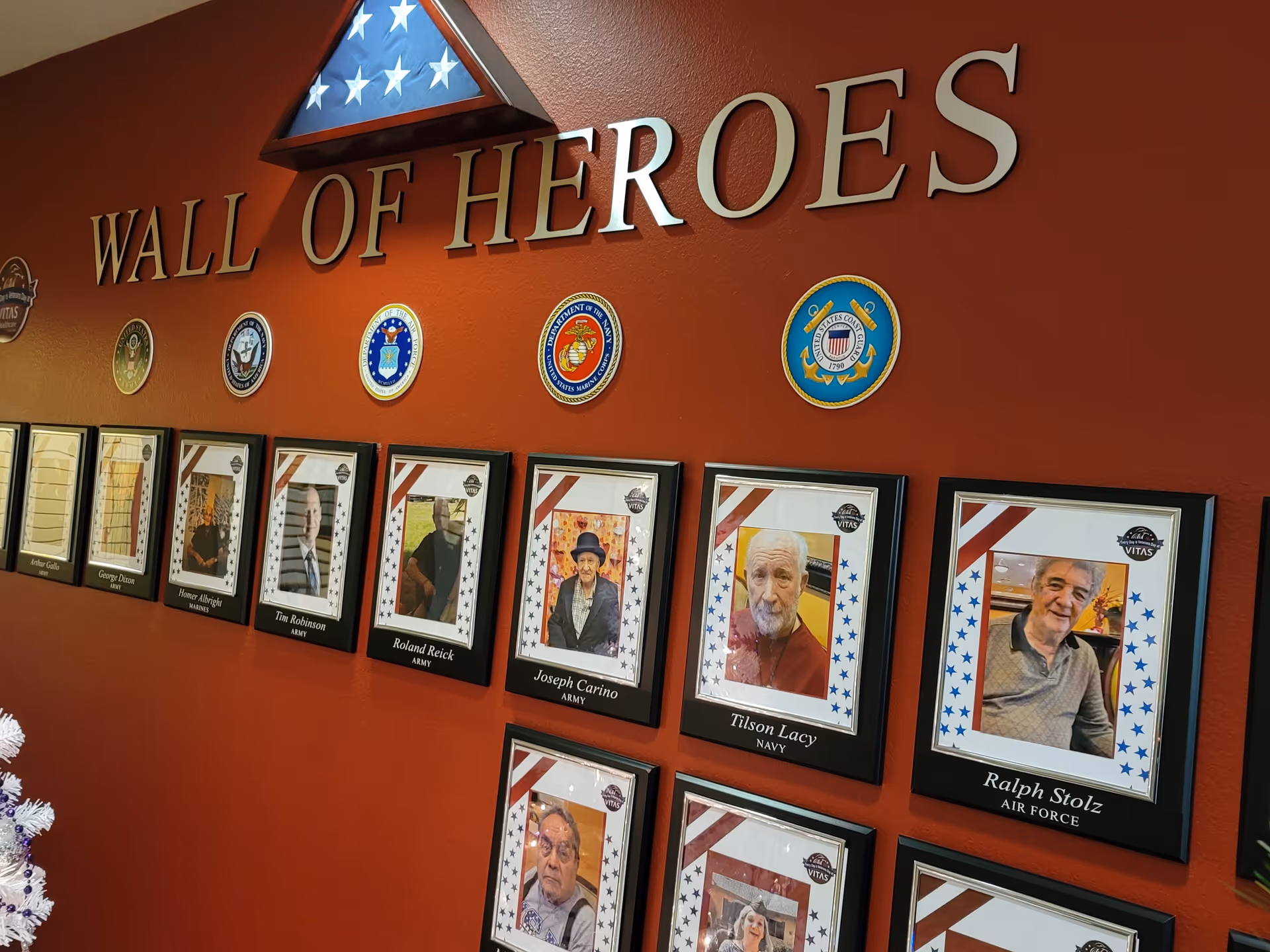 A red wall titled 'Wall of Heroes' displaying framed photographs of senior individuals, each with their name and military branch beneath their picture. Above the photos are emblems representing different branches of the U.S. military, and a folded American flag is mounted at the top center of the wall.