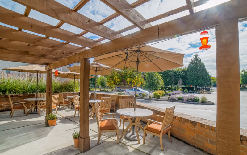 Outdoor patio area with wooden pergola overhead, round tables with wooden chairs featuring orange cushions, large beige umbrellas providing shade, potted plants, hanging flower basket, and a hummingbird feeder. The background shows a parking lot with trees and landscaping under a partly cloudy sky.