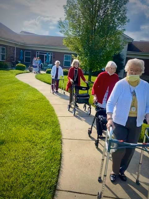 A group of elderly women walking outside on a paved pathway in a garden area of a senior living facility. They are using walkers and are dressed in light sweaters and jackets. The building and green lawn are visible in the background under a partly cloudy sky.