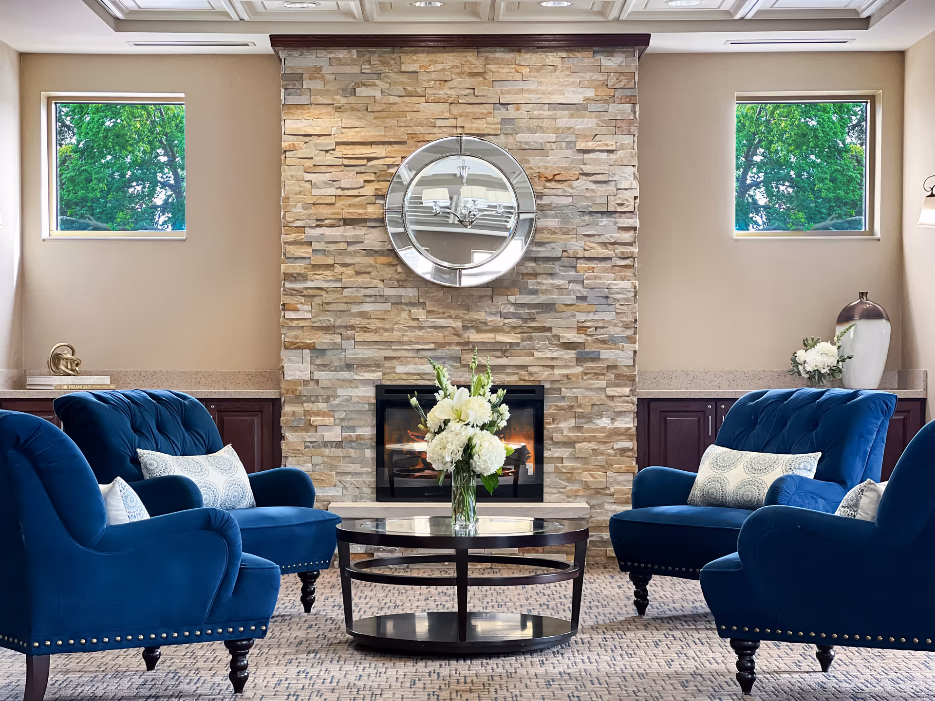 Cozy seating area with four blue armchairs arranged around a round glass coffee table in front of a stone fireplace topped by a round mirror.