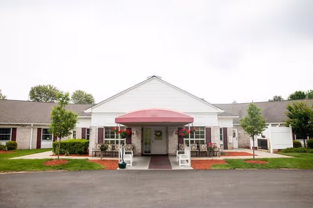 Front entrance of a single-story senior living facility with a red awning, benches, hanging flower baskets, and landscaped grounds.