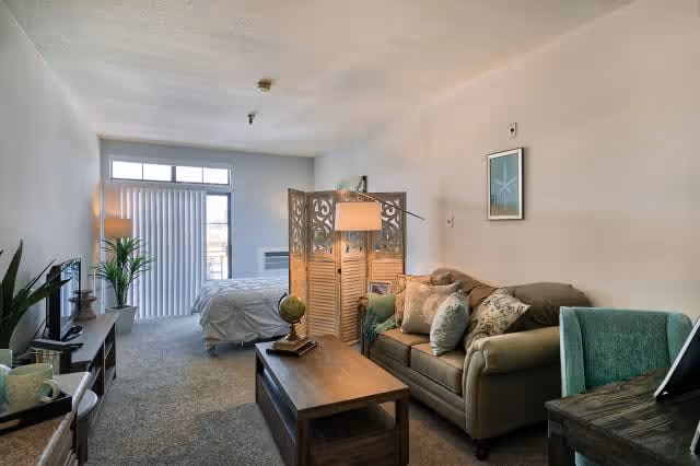 A cozy studio apartment with a living area and sleeping area separated by a decorative wooden room divider. The living area has a green sofa with patterned pillows, a wooden coffee table with a small globe and lamp, and a green upholstered chair next to a small desk. The sleeping area has a bed with white bedding near a window with vertical blinds. The room is carpeted and softly lit with natural light coming through the window.
