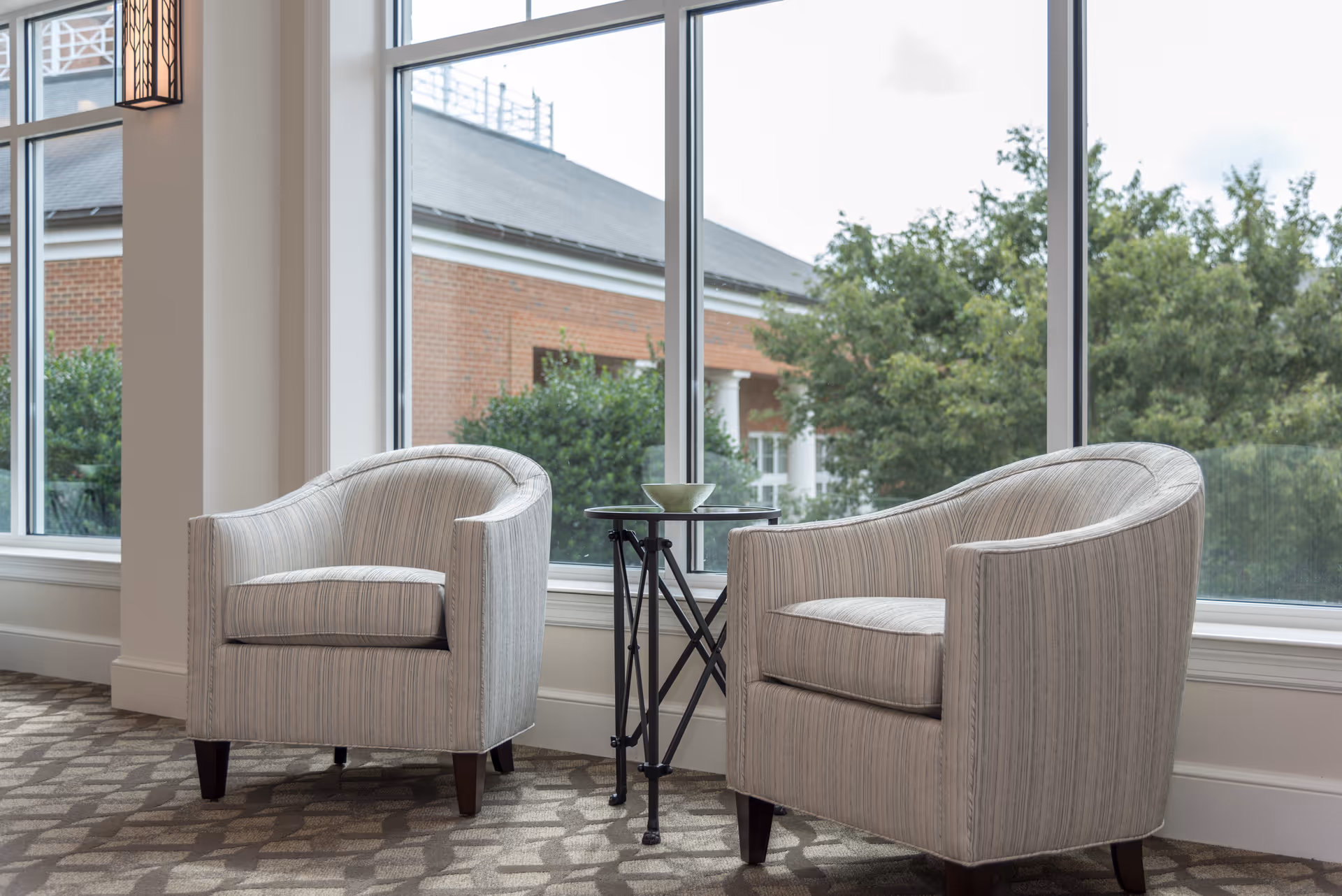 Two light gray upholstered armchairs with subtle vertical stripes positioned near a large window with a view of trees and a brick building outside. A small round black side table with a decorative bowl sits between the chairs on a patterned carpeted floor.