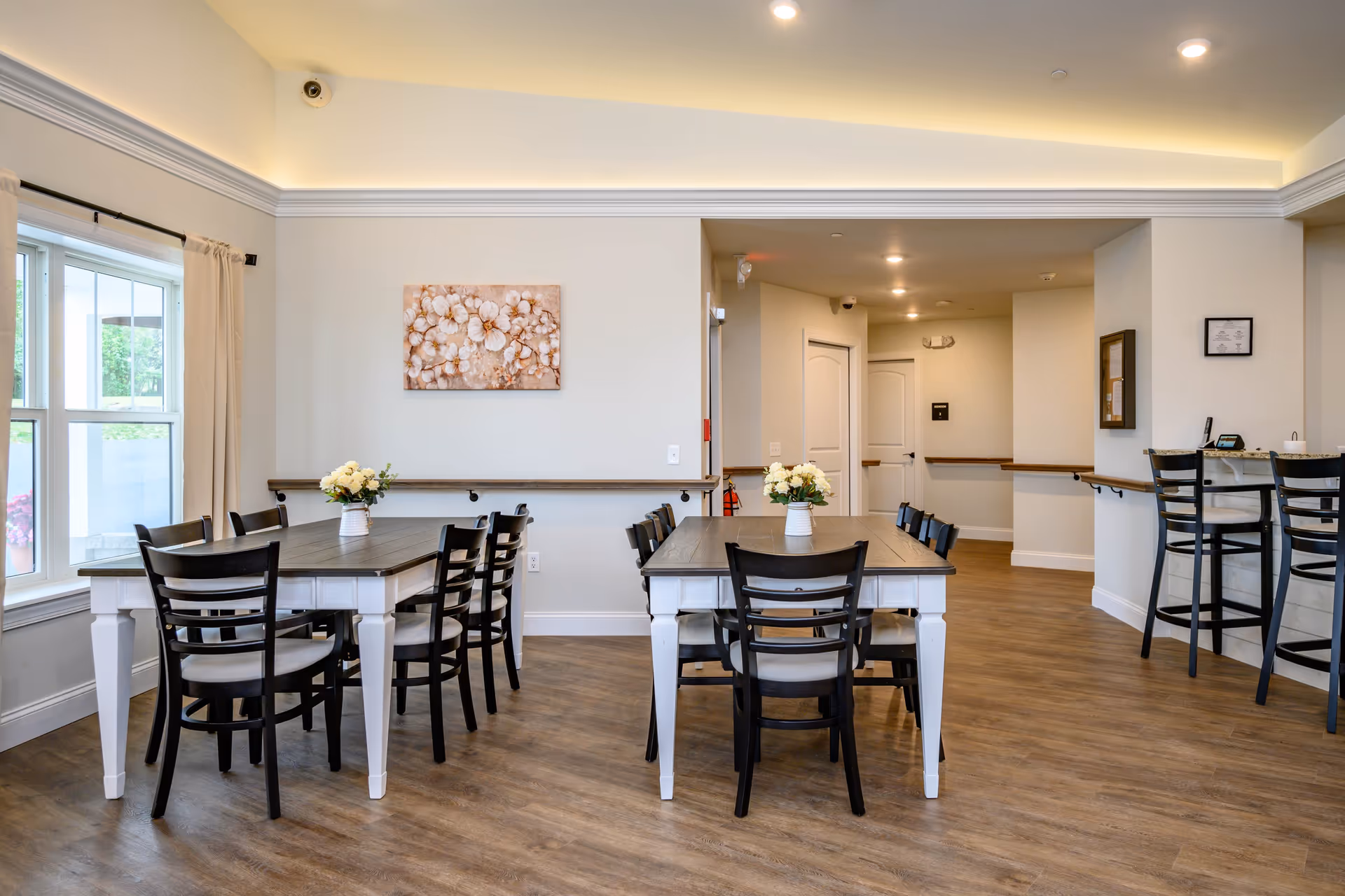 A bright and clean dining area in an assisted living facility with two rectangular tables, each surrounded by black chairs with light-colored cushions. Each table has a vase with white flowers. There is a large window with beige curtains on the left side, and a light wood floor. The walls are light-colored with a floral painting hanging above one table. In the background, there is a small counter with three high chairs and a hallway with white doors.