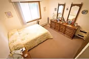 A bedroom with a bed covered in a yellow floral bedspread, a wooden dresser with two mirrors, various decorative items on the dresser, a window with curtains, and a small side table with a lamp.