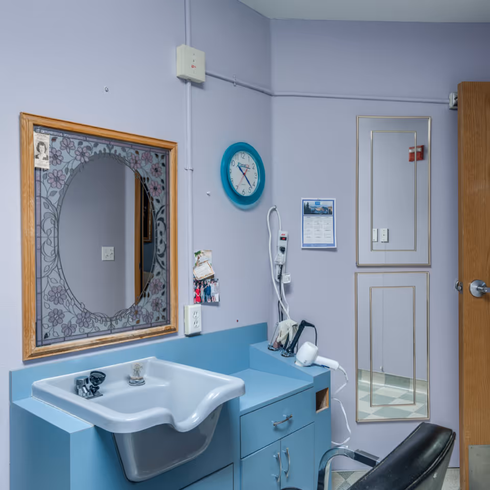 Interior view of a hair washing station in a senior living facility. The station includes a blue sink with a contoured neck rest, a mirror with a floral design frame, a blue clock on the wall, a calendar, a hair dryer, and a black salon chair. The walls are painted light purple and there is a door with a mirror attached.