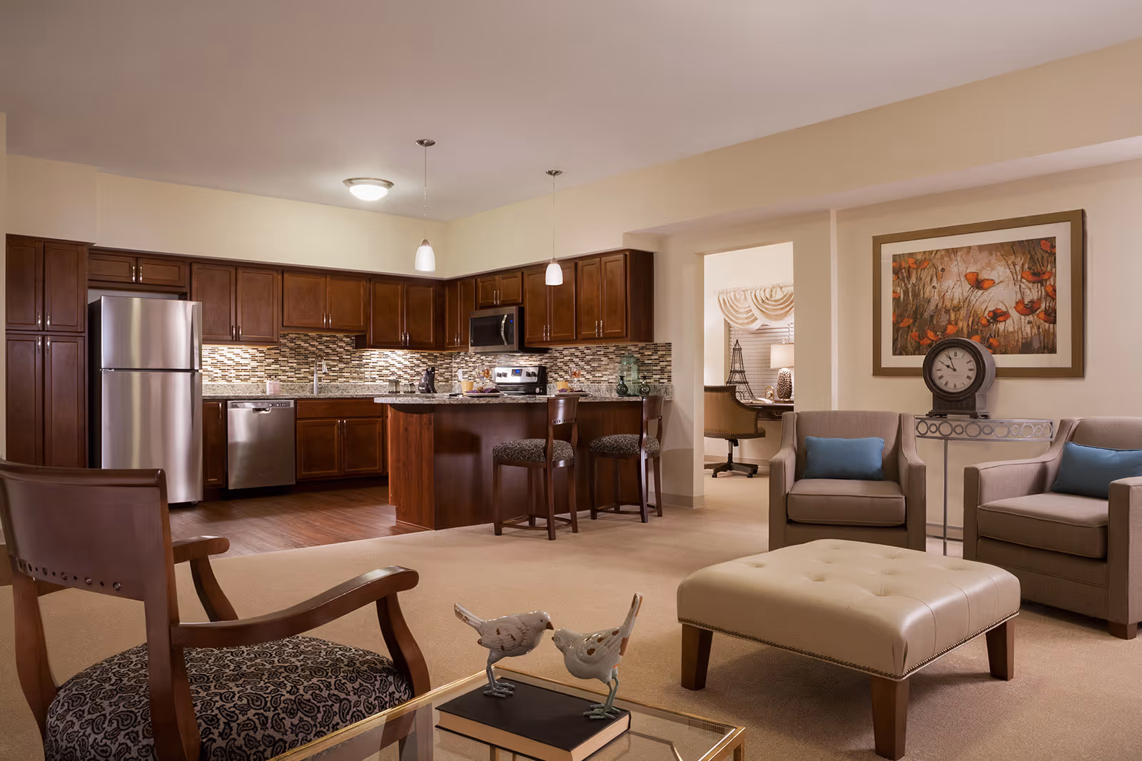 A cozy open-concept living area and kitchen in a senior living facility. The kitchen features dark wood cabinets, stainless steel appliances, a tiled backsplash, and a granite countertop island with two bar stools. The living area has two beige armchairs with blue cushions, a beige ottoman, a glass coffee table with decorative bird figurines, and a framed floral painting on the wall. A small office space with a desk and chair is visible through an open doorway.