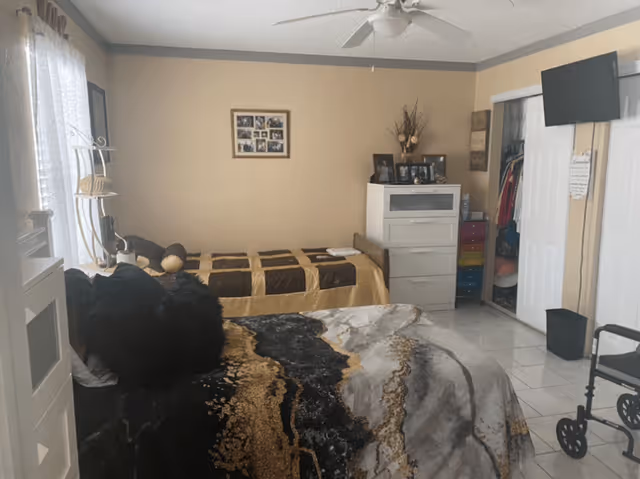 A bedroom with two beds, one in the foreground with a black, gold, and gray patterned comforter and black pillows, and another twin bed in the background with a brown and beige checkered comforter. The room has beige walls, a ceiling fan, a white dresser with framed photos and decorative items on top, a closet with sliding doors, a wall-mounted TV, and a black walker near the door. The floor is tiled in white.