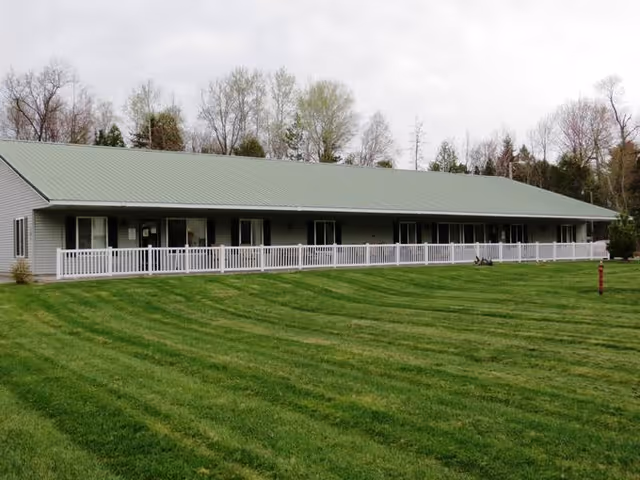 Single-story building with a green metal roof and light gray siding, featuring multiple windows and a white railing along the front porch. The building is surrounded by a well-maintained green lawn and trees in the background under a cloudy sky.