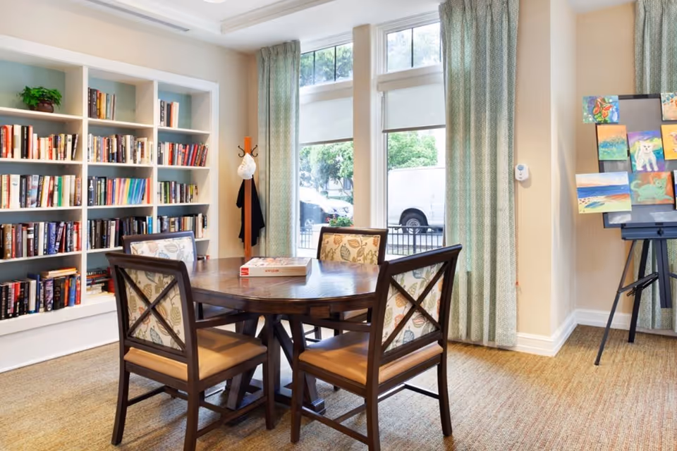 A cozy room with a round wooden table surrounded by four chairs with floral patterned cushions. Behind the table is a built-in bookshelf filled with books. Large windows with light green curtains allow natural light to brighten the room. To the right, an easel displays colorful paintings.
