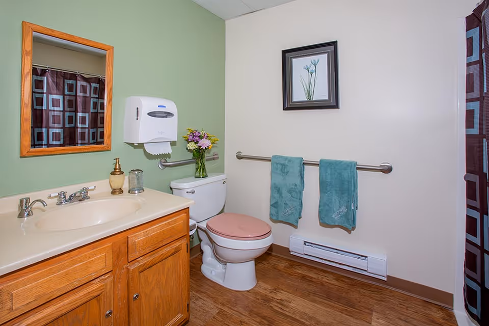 A bathroom with a wooden vanity and sink, a mirror with a wooden frame, a toilet with a pink seat, a paper towel dispenser, a vase with flowers on the toilet tank, two teal towels hanging on a metal grab bar, a framed flower picture on the wall, and a shower curtain with a geometric pattern.