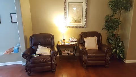 Two brown leather armchairs flank a small side table with a lamp and magazines against a beige wall, with framed artwork above and a potted plant to the right.