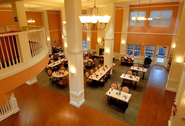 Two-story dining room with multiple set tables, chandeliers, large columns and an upper balcony in a retirement facility.