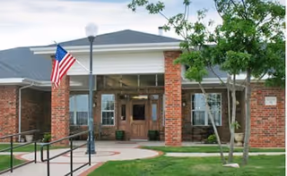 Exterior view of a brick building with a covered entrance supported by white columns, an American flag on a pole, a street lamp, and a ramp leading to the entrance. There are trees and green grass around the walkway.