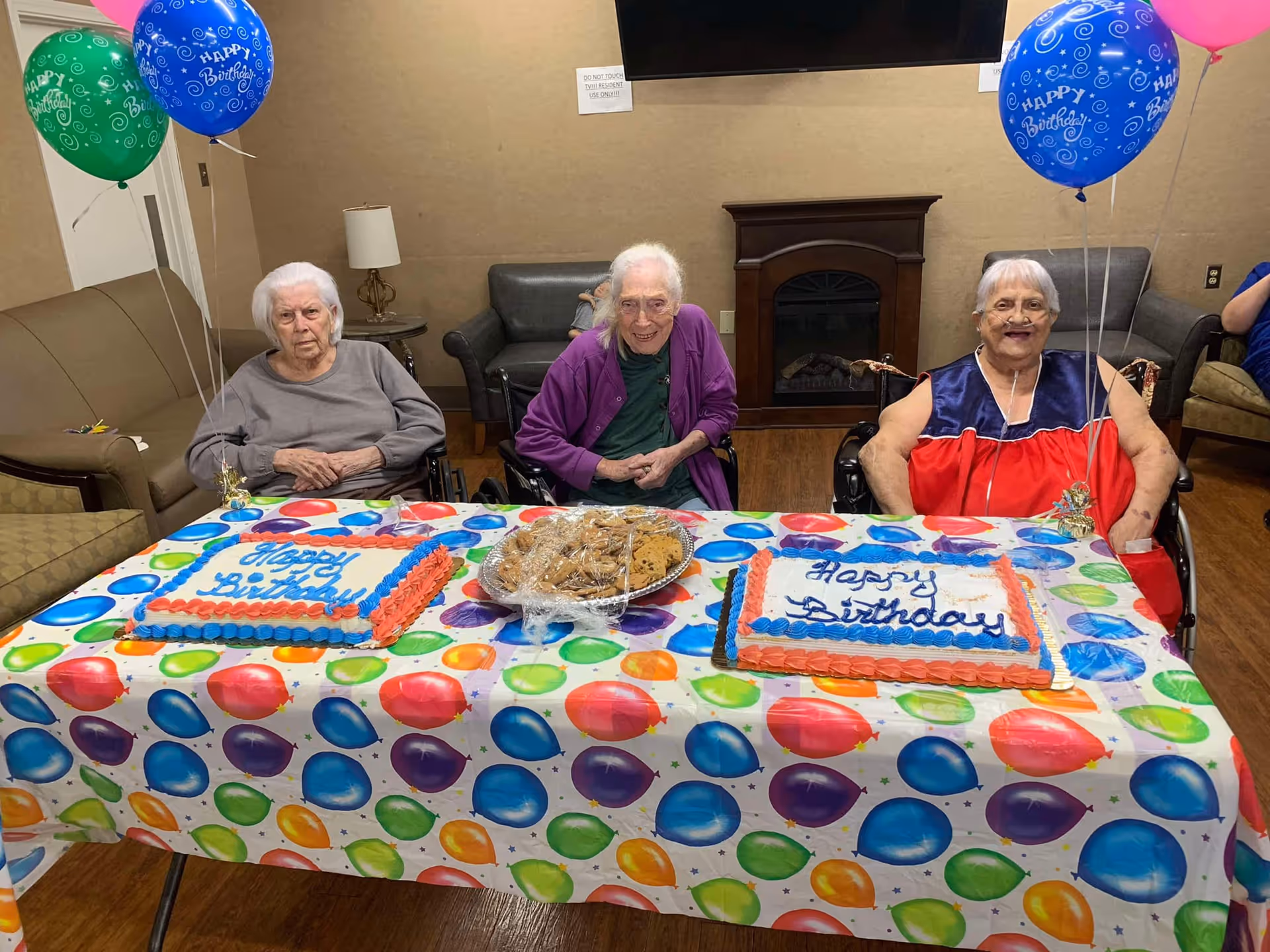 Three elderly women sitting at a table decorated with a colorful balloon-patterned tablecloth. Each woman is in a wheelchair, and there are two birthday cakes and a plate of cookies on the table. The room has a cozy interior with a fireplace, a TV mounted on the wall, and balloons that say 'Happy Birthday' tied to the table.