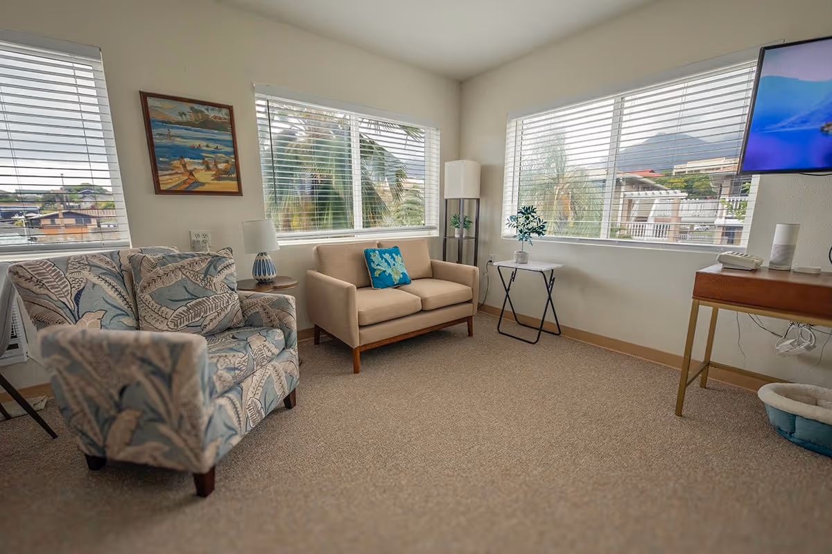 A bright living room with large windows showing an outdoor view with palm trees and mountains. The room contains a patterned armchair, a beige loveseat with a blue decorative pillow, a small side table with a lamp, a tall floor lamp, a small table with a plant, and a wall-mounted TV. The carpet is beige and the walls are light-colored.