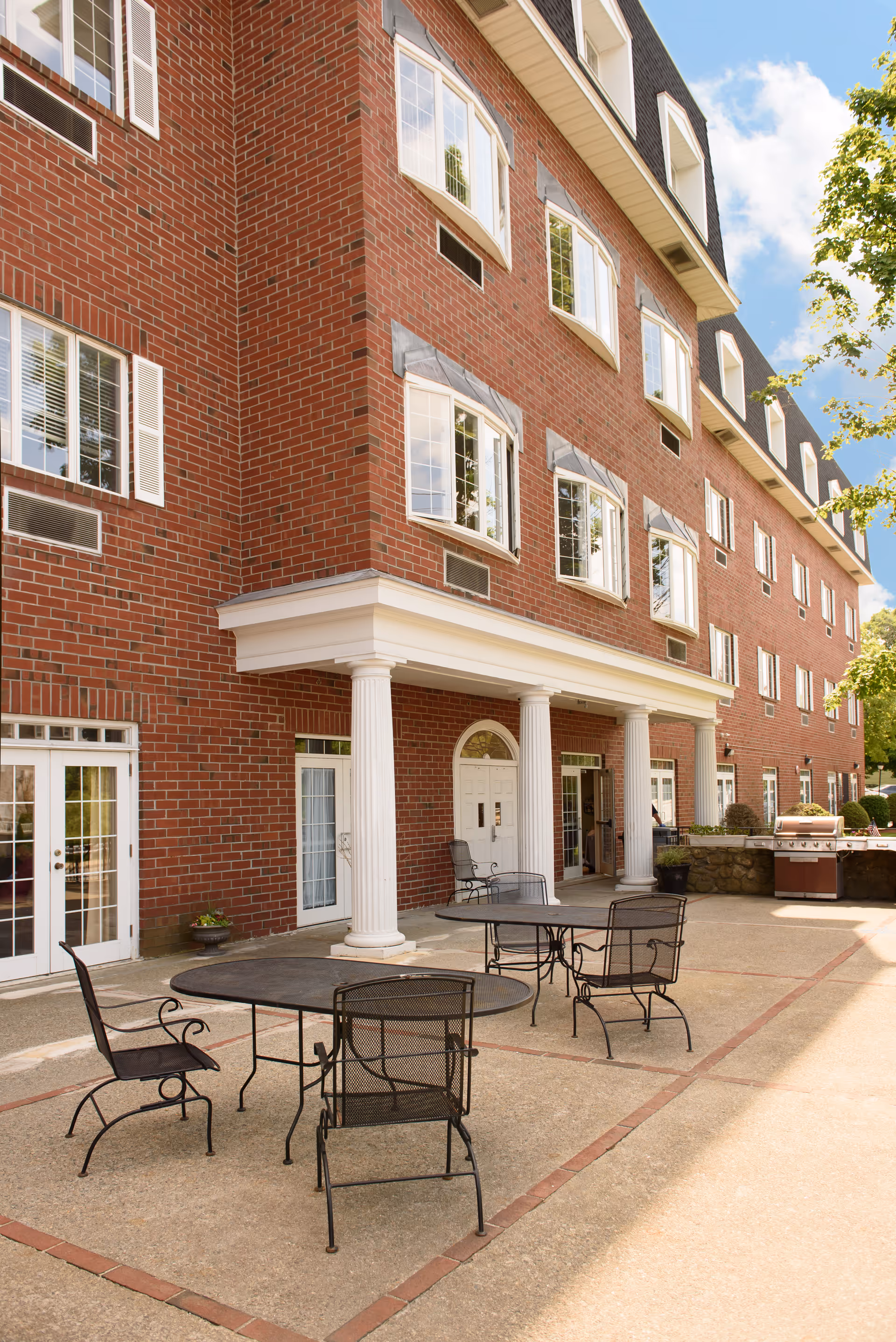 Outdoor patio with metal tables and chairs in front of a red brick nursing facility featuring white columns and multiple windows.