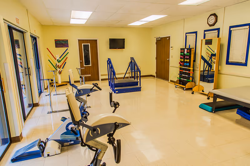 A rehabilitation room with exercise equipment including stationary bikes, handrails for walking practice, a rack of colorful dumbbells, a large mirror, and a therapy table. The room has beige walls, a tiled floor, and several doors and windows with blinds.