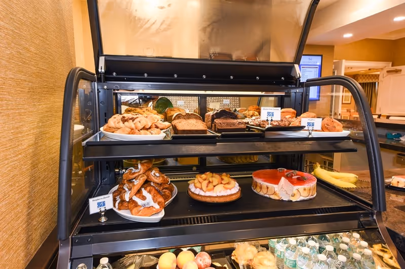 A bakery display case filled with various pastries, breads, and desserts including blueberry danish, fruit tart, and a red gelatin dessert. Bottled water and fresh fruit are visible on the lower shelf. The setting appears to be a food service area within a senior living facility.