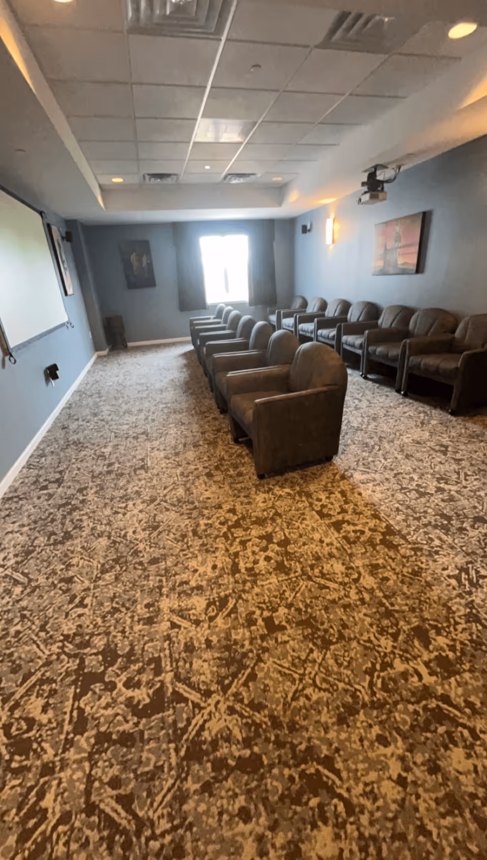 A small theater room with two rows of brown cushioned armchairs facing a large white projection screen on the left wall. The room has patterned carpet flooring, blue-gray walls, a window with curtains at the far end, and framed artwork on the walls. A ceiling-mounted projector is visible near the front of the room.