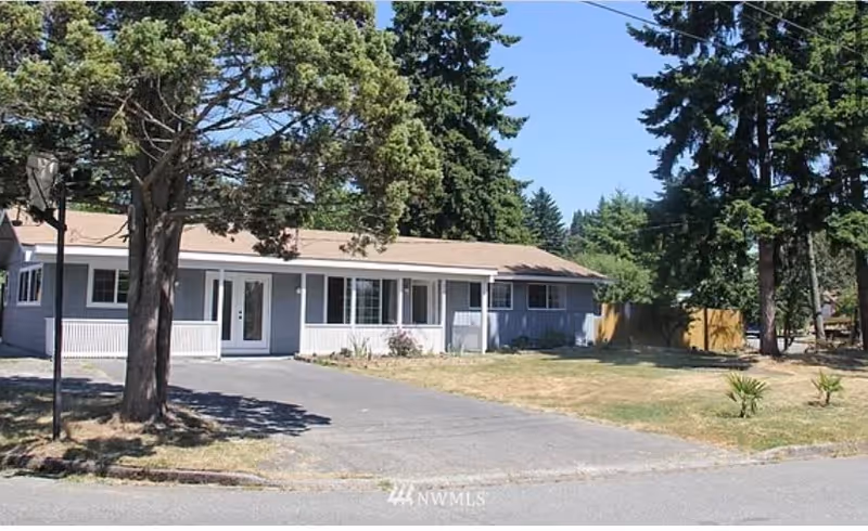 Single-story residential building with a light gray exterior and white trim, surrounded by tall trees and a lawn with some dry patches. The building has a covered porch area and a paved driveway leading up to it under a clear blue sky.