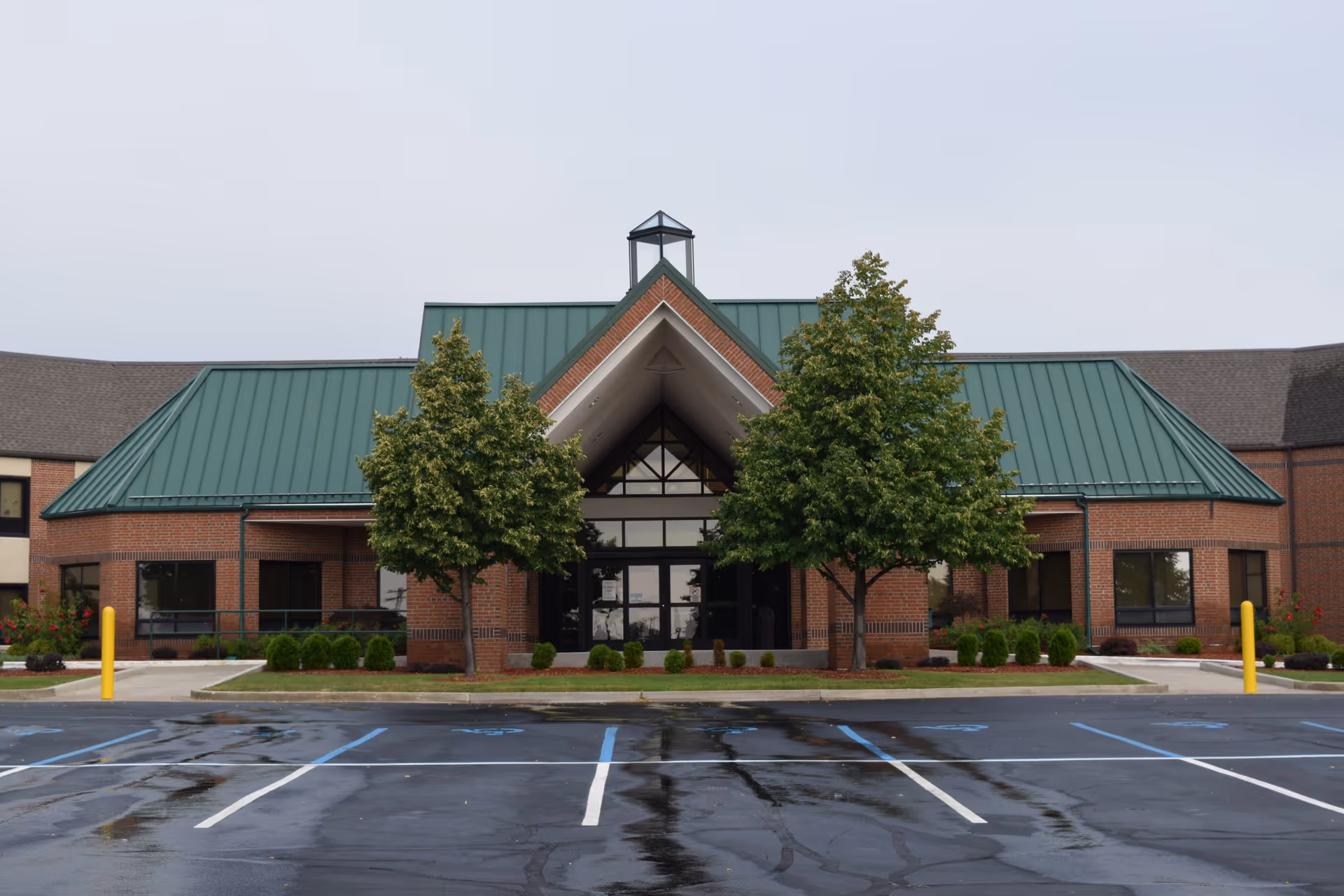Front exterior view of Tuscola County Medical Care Community building with a green metal roof, brick walls, two trees flanking the entrance, and an empty parking lot with wet pavement.